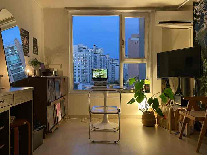 ホワイトとウッドトーンが調和するモダンなワンルーム、透明な折りたたみ椅子と丸テーブル、テレビと植物が配置された落ち着いたホームオフィスのインテリア