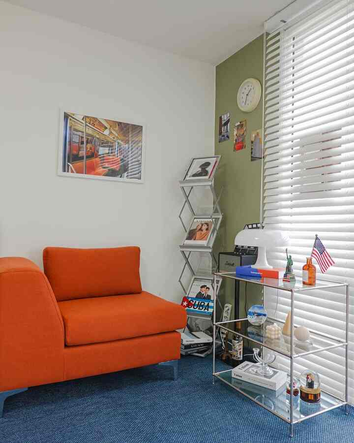 グリーンのアクセントウォールとオレンジのソファ、ブルーのラグが配置された寝室で、ポスターやインテリアオブジェが印象的な空間です。