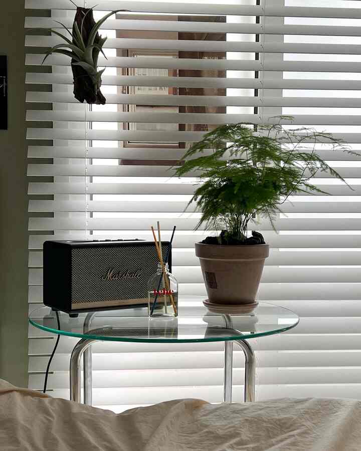 白いウッドブラインドを背景に、中央に透明なガラステーブル、緑の植物の鉢、マーシャルスピーカー、ディフューザーが配置されたナチュラルな雰囲気のリビングルーム