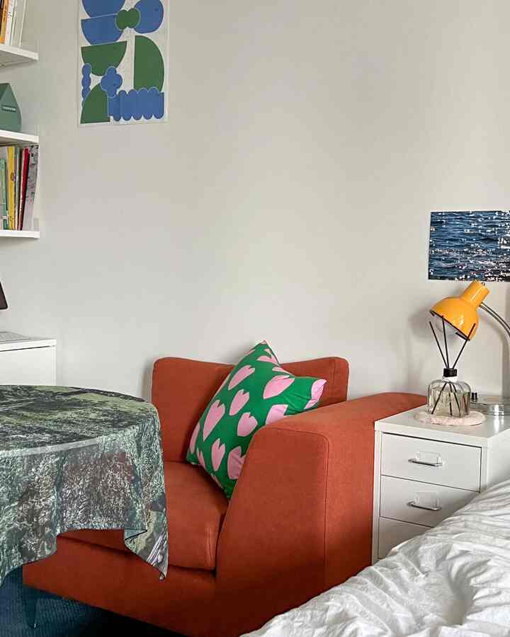 白い壁とオレンジ色のソファ、緑とピンクのクッション、横に白いチェストと黄色いテーブルランプが配置されたコンパクトな寝室