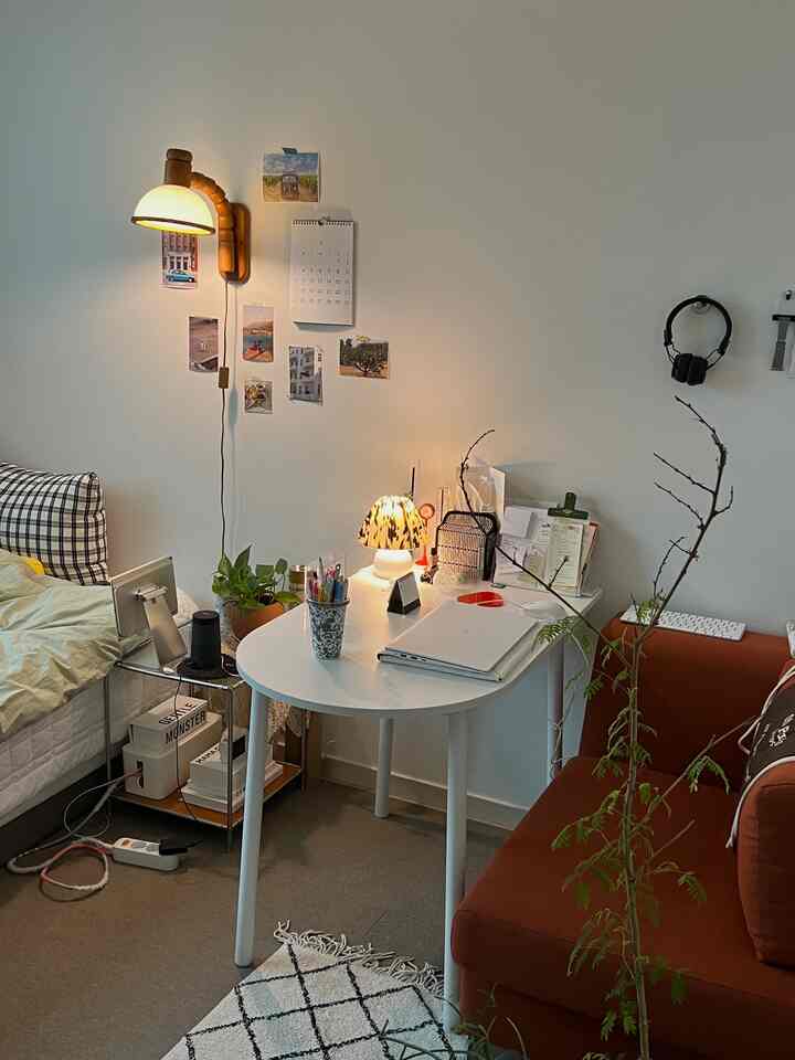 白い壁と木目調の壁灯がある一人暮らしの寝室兼ホームオフィススペース、白いデスクとオレンジ色のソファが調和した雰囲気のあるインテリア