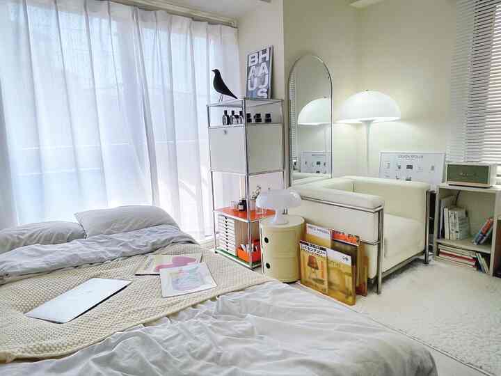 明るいモノトーンのワンルーム寝室、清潔感のある白い寝具とシアーカーテン、クリーム色のソファとフロアランプが配置された落ち着いた雰囲気のインテリア