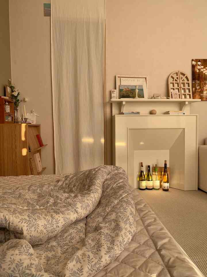 ベージュとホワイトの色調の寝室、居心地の良い寝具と暖炉の棚が配置された温かみのある朝の雰囲気のあるインテリア