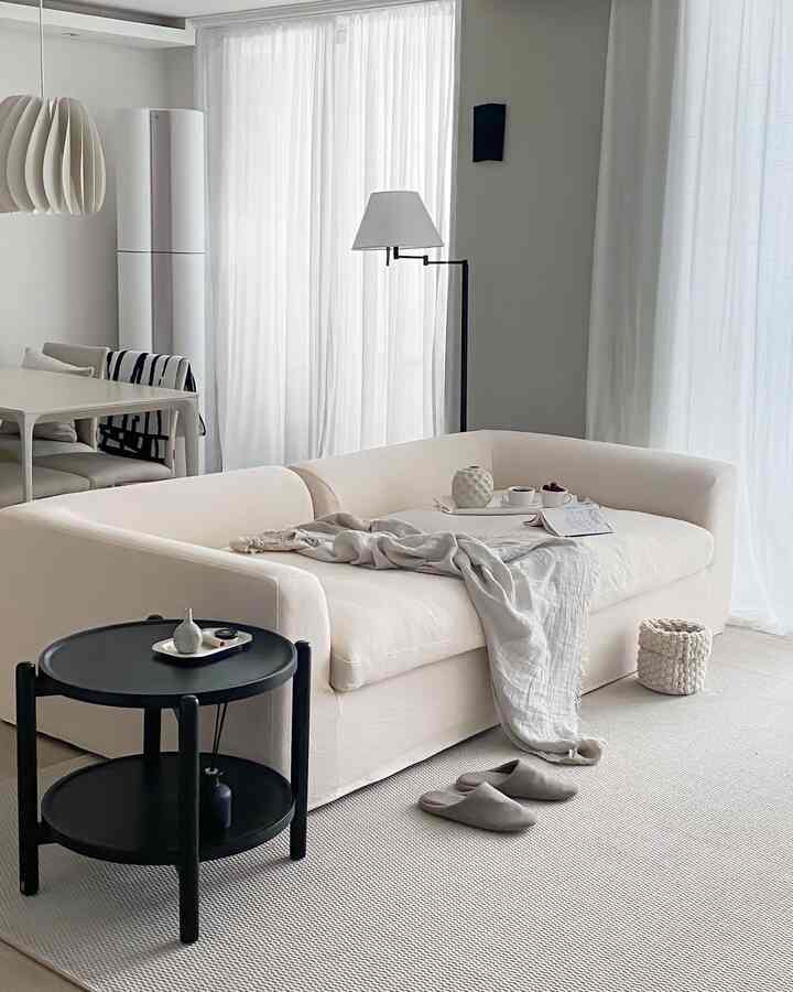 白いトーンのリビングルーム、クリーム色のソファと黒の丸テーブル、明るいカーテンが配置されたシンプルでナチュラルモダンな雰囲気のインテリア