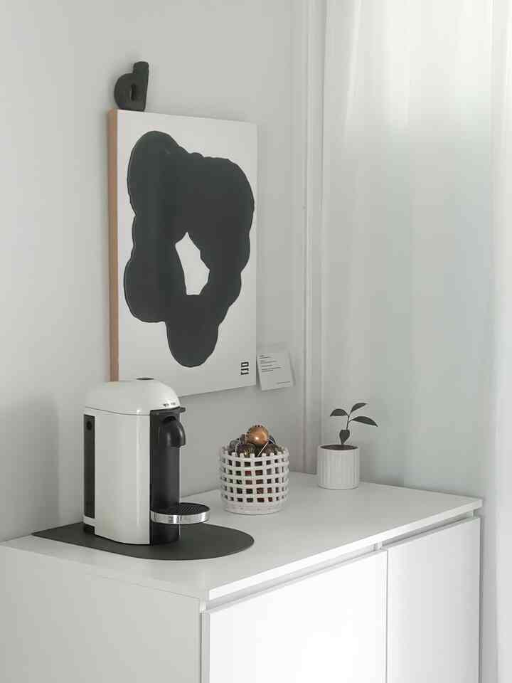 白を基調としたキッチンスペースに、コーヒーマシンとカプセルホルダー、抽象画の額縁が配置されたミニホームカフェコーナー