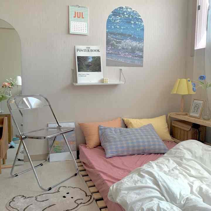 ナチュラルカラーの小さな寝室、木製ベッドフレームにピンクの寝具とカラフルなクッション、透明な折りたたみ椅子が配置された清潔感のある空間