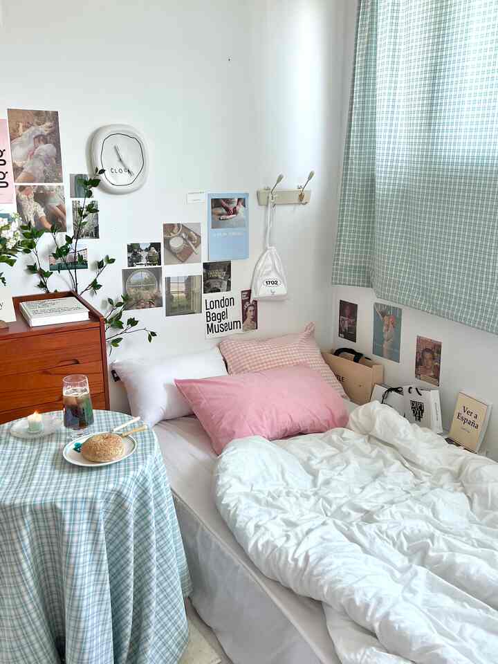 ホワイトとパステル調の一人暮らし寝室、ヴィンテージポスターとチェック柄カーテン、かわいらしいクッションと植物が配置された雰囲気のあるインテリア
