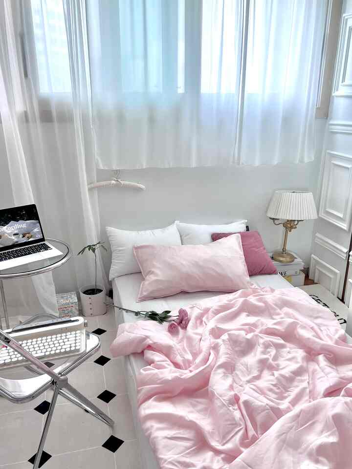 柔らかなホワイトトーンとピンクの寝具が調和する寝室、透明チェアと丸いサイドテーブルが配置された空間