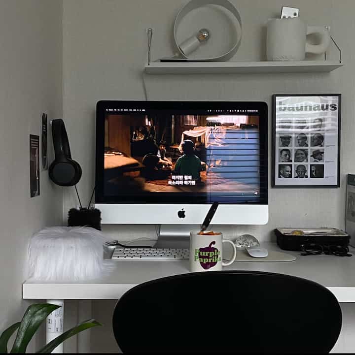 白を基調としたコンパクトなホームオフィス空間、iMacのデスクセットアップとヘッドホン、額縁のポスターとミニマルな照明が配置されたインテリア