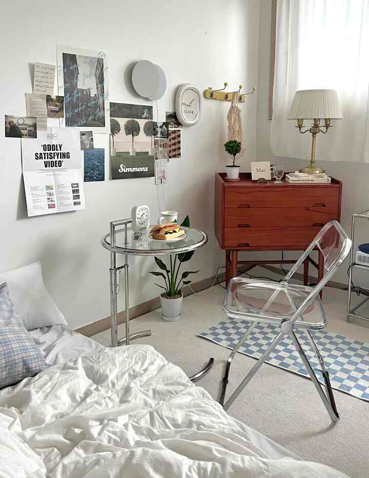 白とウッドトーンのミニマルな寝室、木製チェストと透明な椅子、整った寝具が配置された印象的な空間