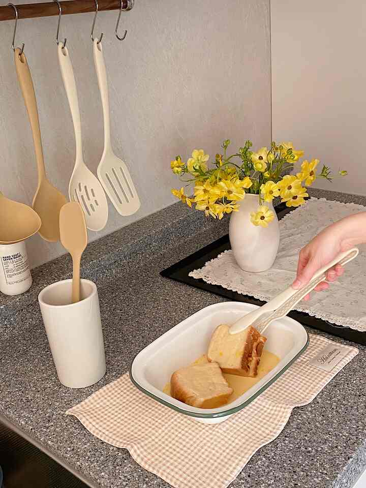 ベージュとクリーム色のシリコン調理器具が掛けられ、黄色い花の飾りとパンが入った器が置かれた、落ち着いた雰囲気のキッチン