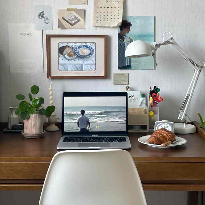 ホワイトの壁と木製デスクが調和したホームオフィス、ノートパソコンとデスクランプ、キャンドル、額縁が配置された落ち着いた作業空間