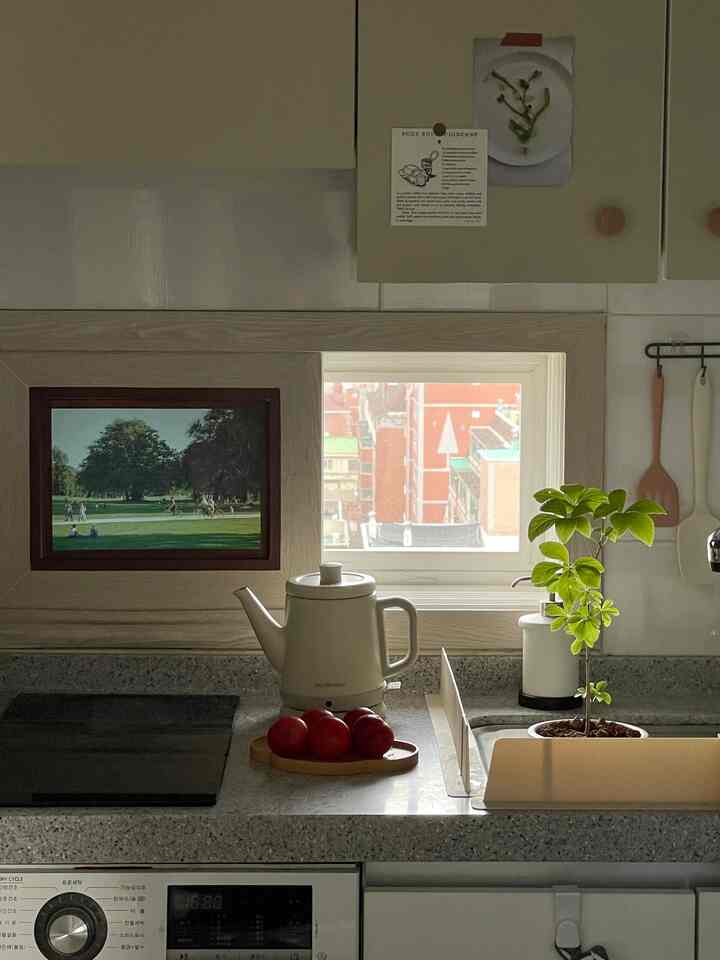 白と緑のトーンのキッチンで、窓の隣にポスターと電気ケトルがあり、シンクのそばに植物が置かれた雰囲気のあるインテリア