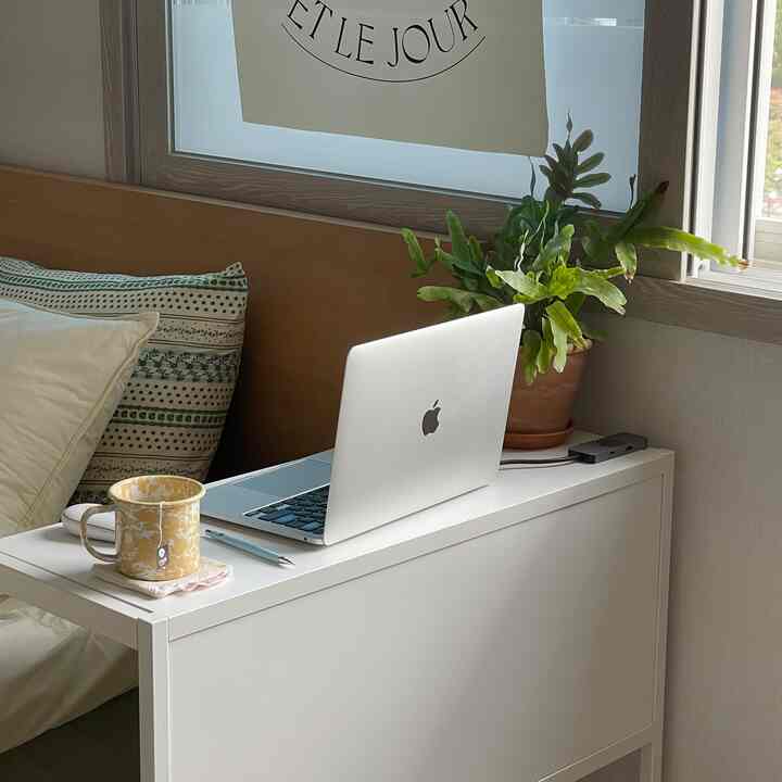白と木目調が調和する寝室、ベッドサイドの簡易作業台にノートパソコンとマグカップ、窓際の緑色の植物が明るい雰囲気を演出