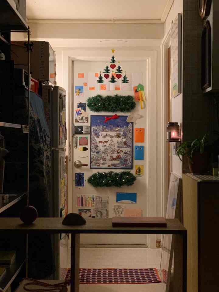 暖かい照明の下、狭い廊下にある白い玄関ドアにクリスマスの飾りや多彩なポスターが配置された雰囲気のある空間