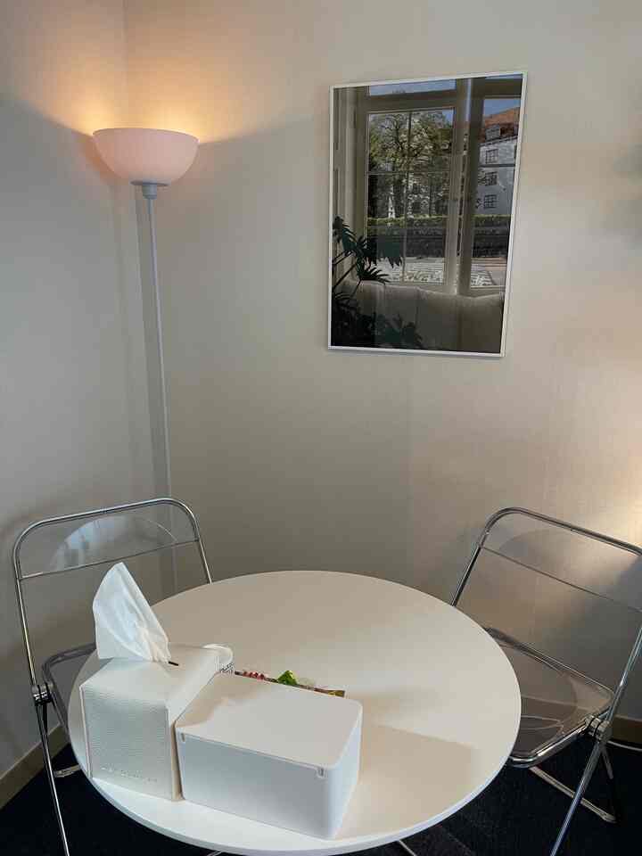 明るいベージュ調の狭い部屋のコーナーにラウンドホワイトテーブルと透明な折りたたみ椅子2脚、壁には窓の映り込みがある額縁が配されたおうちカフェ