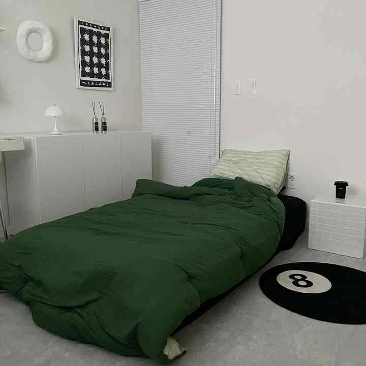 白を基調としたモダンな寝室、ダークグリーンの布団と枕が置かれたベッドが中央にあり、白いブラインドとナイトテーブルが配置されたシンプルなインテリア