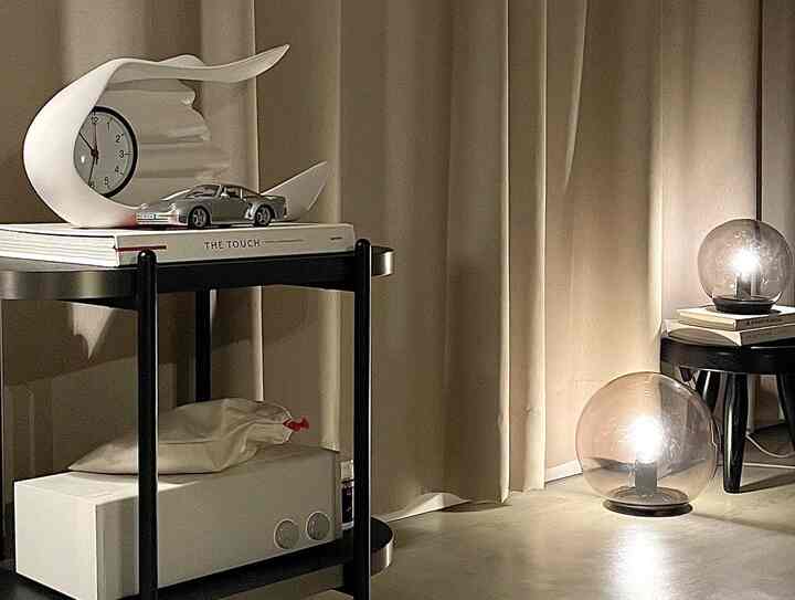 ベージュの遮光カーテンと黒いナイトテーブル、透明なムードライトが配置された安らぎのある寝室の一角