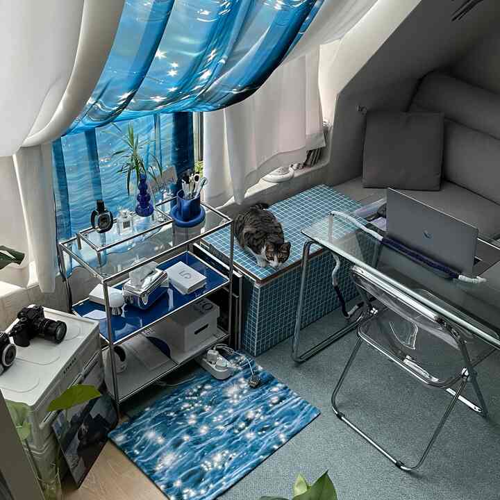 青い波模様のカーテンとタイルスツールがあるコンパクトなホームオフィス、猫が座り、グレーのソファと透明なデスクが配置された空間