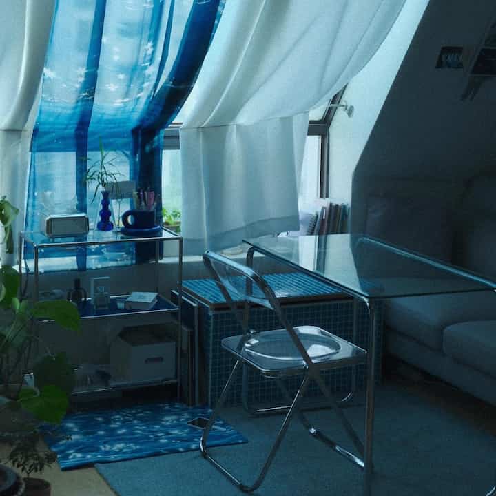 ホワイトとブルーの色調の小さなリビングスペース、透明な折りたたみ椅子とガラステーブル、遮光カーテンが配置された雰囲気のあるインテリア