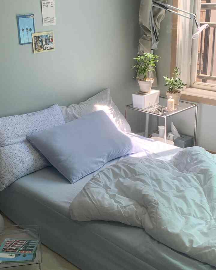 柔らかなナチュラルカラーの壁と窓がある寝室、ホワイトとライトブルーの寝具、ナイトテーブルと植物が配置された落ち着いた空間