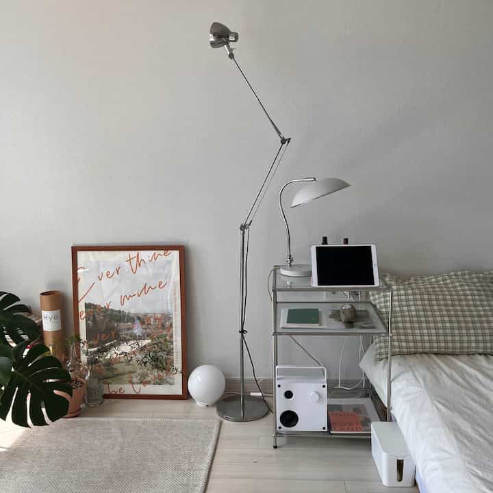 明るい白色調の寝室、チェック柄の枕と金属製の収納棚、フロアランプが調和したシンプルで北欧風のインテリア