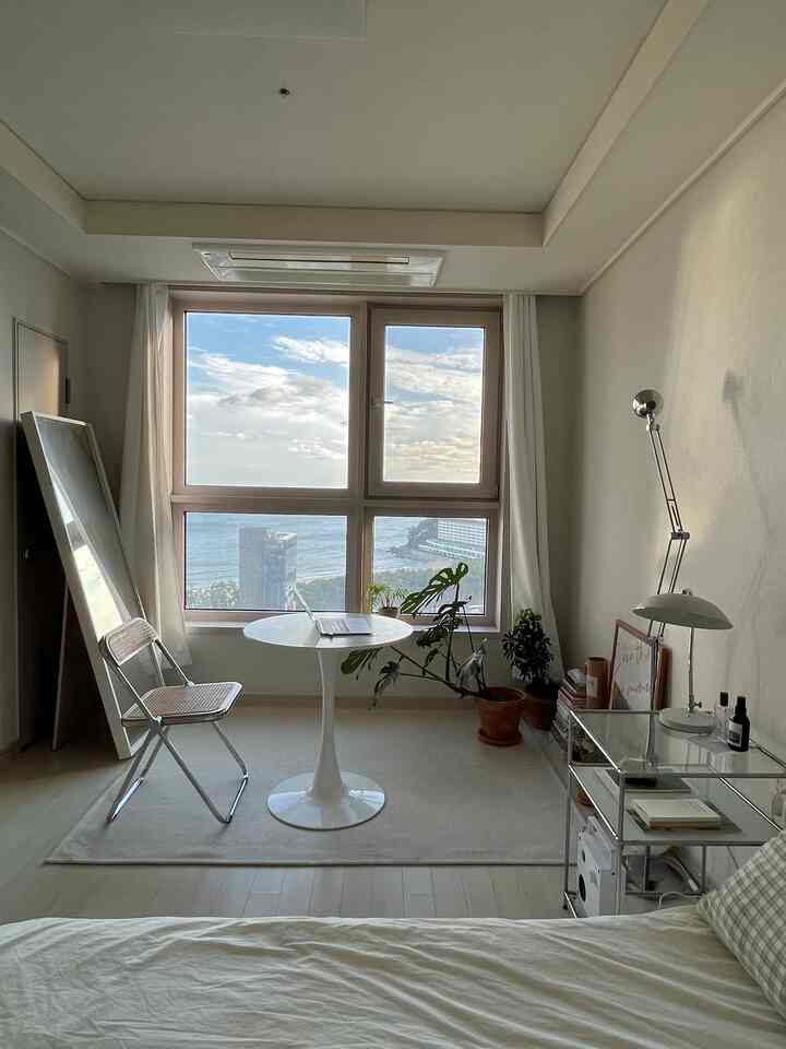 明るいホワイトと木目調の調和がとれた小さな6畳の寝室兼ホームオフィス、窓際に丸テーブルと植物が配置されたすっきりとした空間