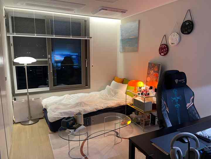 白とネイビーのトーンが特徴のワンルーム寝室、ベッドとゲーミングチェア、モダンな照明と透明なガラステーブルが配置された居心地の良いインテリア