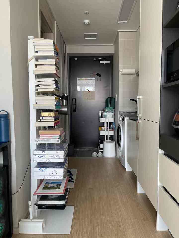 白と木目調が調和したワンルームの玄関とキッチン、棚と収納カートが効率的に配置されたコンパクトで清潔感のある空間