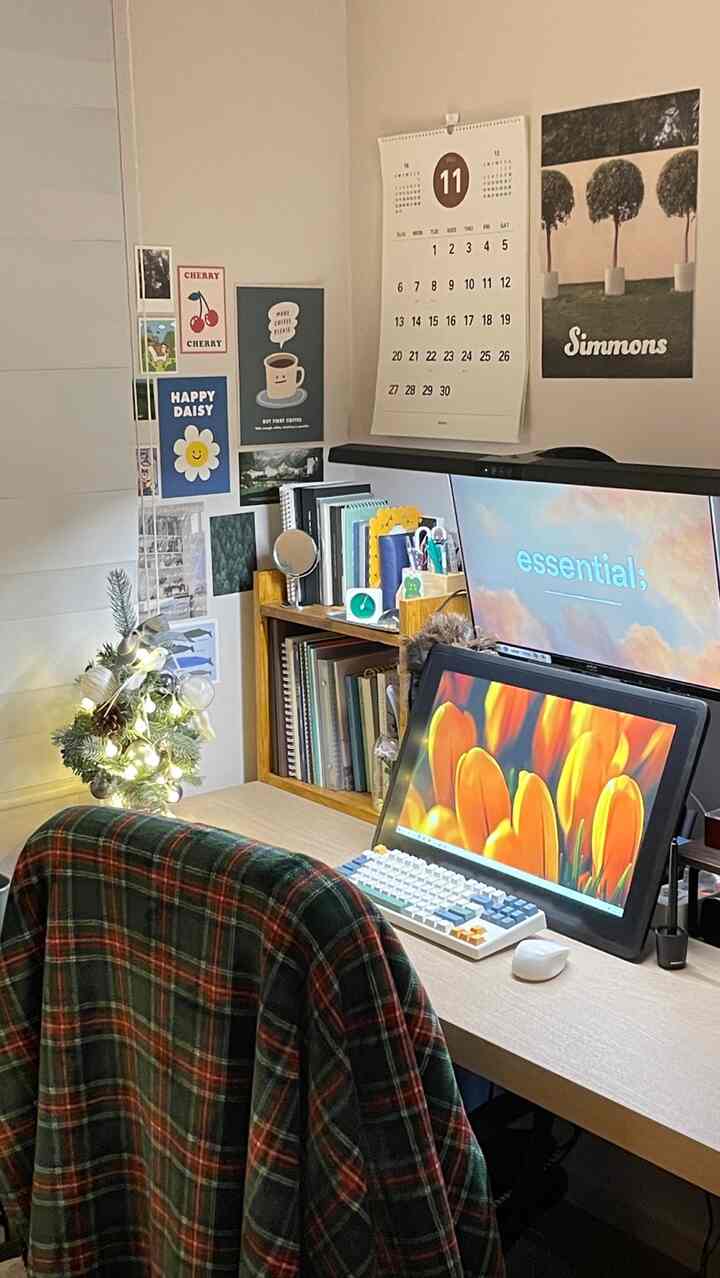 白い壁と木製デスクを備えたコンパクトなホームオフィス、書棚とクリスマスのミニツリー、ポスターが飾られた落ち着いた空間
