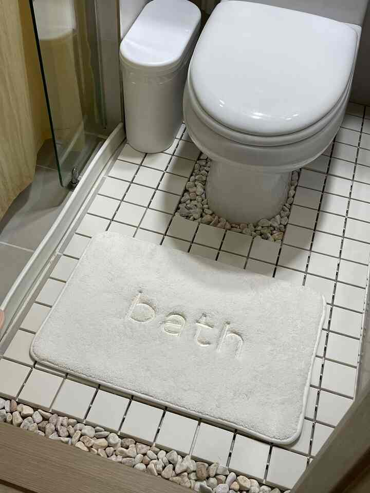 白いタイルの小さなトイレ、トイレ本体、ゴミ箱、バスマットが配置された清潔感のある空間