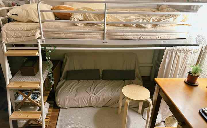 白とベージュの色調のワンルーム、ロフトベッドの下にベージュのソファベッドと木製スツール、自然光が差し込む温かみのあるインテリア