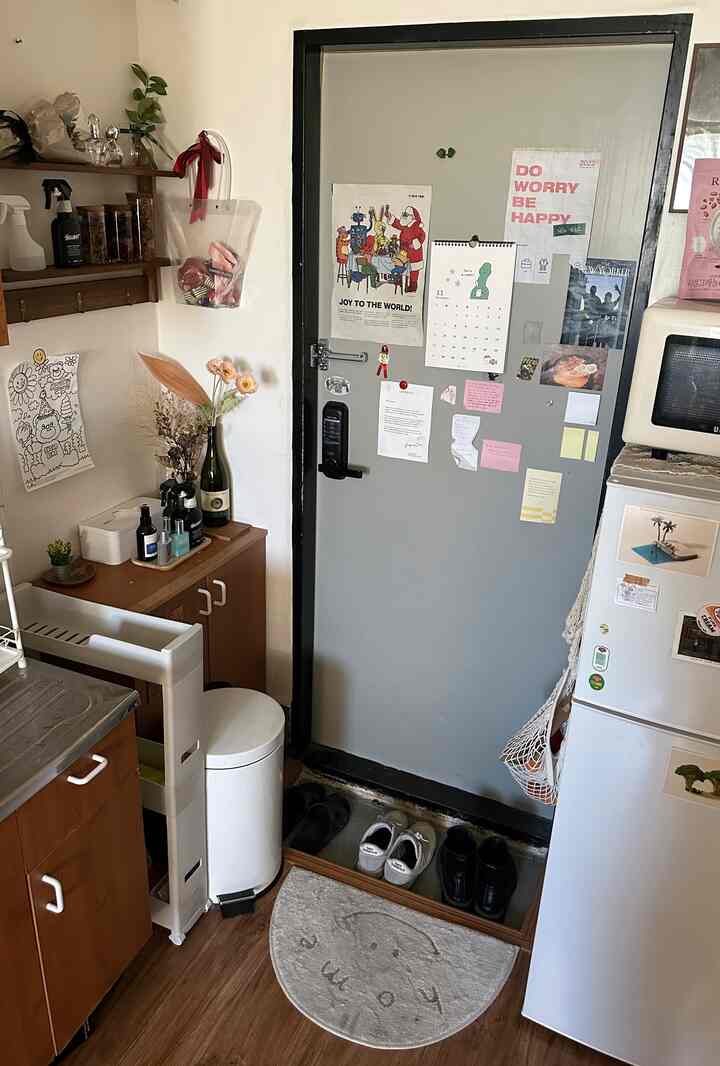 ブラウンの木目調フローリングとホワイトのペダル付きゴミ箱、ポスターが貼られたグレーの玄関扉があるコンパクトな玄関スペース