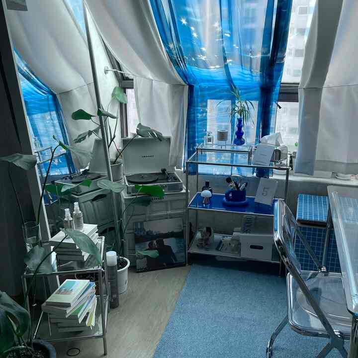 白と青のトーンの多目的空間、透明な椅子とヴィンテージターンテーブル、植物とミニマルな棚が調和した雰囲気のあるインテリア