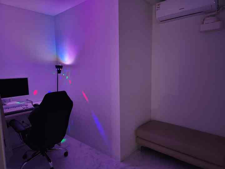 白い壁と床のコンパクトなホームオフィス、黒いオフィスチェアとデスクが配置され、フロアランプからの多彩な光が雰囲気を演出する印象的な空間