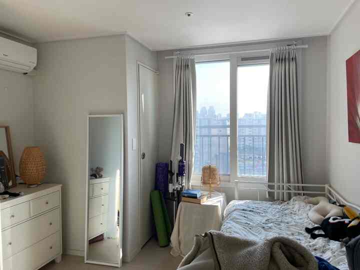 ホワイトとベージュのトーンのワンルーム寝室、鉄製ベッド、チェスト、ラタン照明、遮光カーテンが配置された雰囲気のあるインテリア