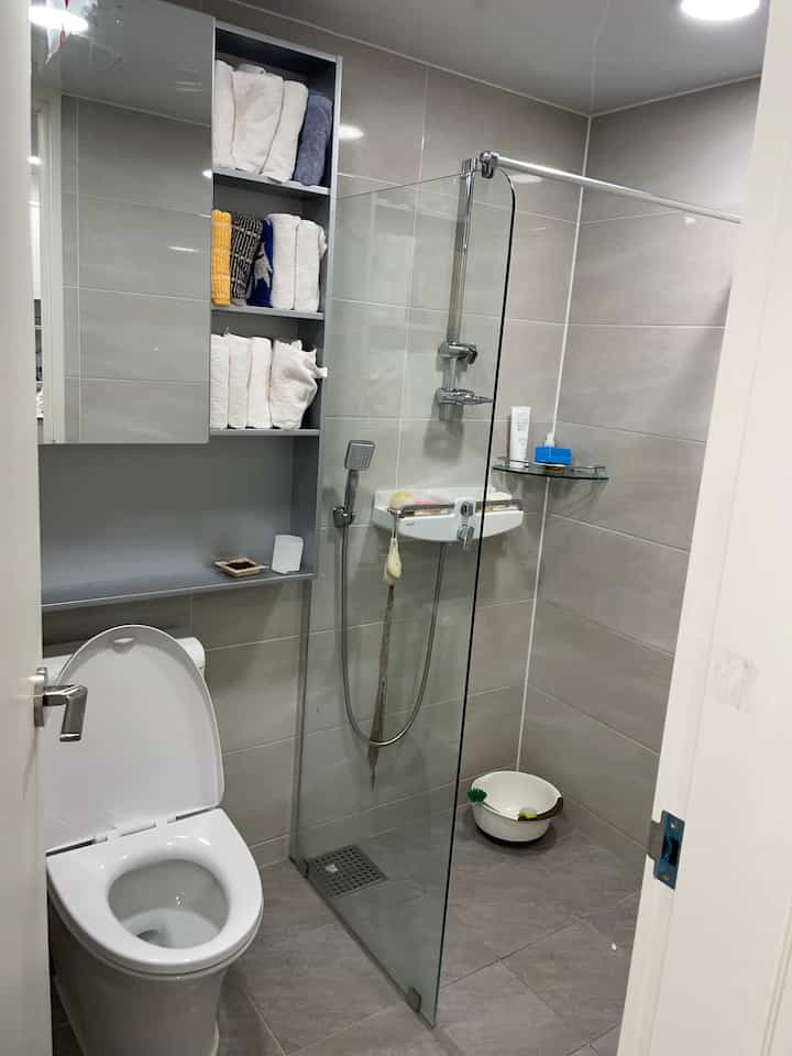 グレーのタイルが施された狭く分離されたバスルーム、トイレとシャワーはガラスの仕切りで区切られ、タオルが棚に整頓された清潔な空間