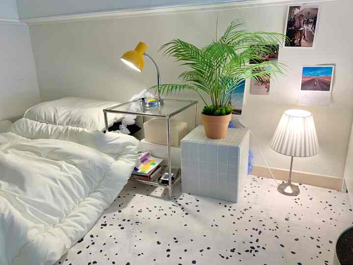 白と緑の色調の狭い寝室、白いラグと金属のナイトテーブル、黄色のスタンドランプが配置されたシンプルな空間