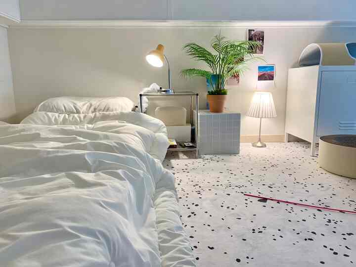 白とナチュラルな色調の寝室、白いベッドとナイトテーブル、ラグ、フロアランプ、植物が配置された落ち着いた空間