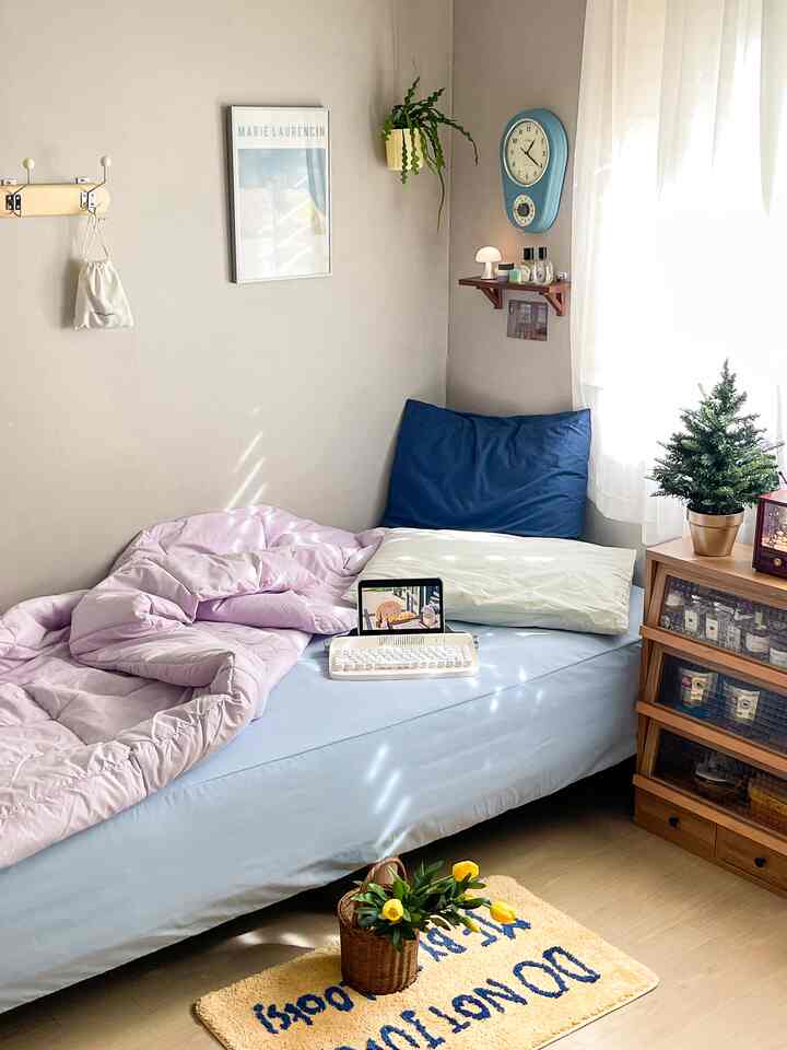 ベージュの壁と木目調のチェストがある6畳寝室、淡いブルーのベッドとクッション、ラベンダー色の布団が自然光に包まれた落ち着いた空間