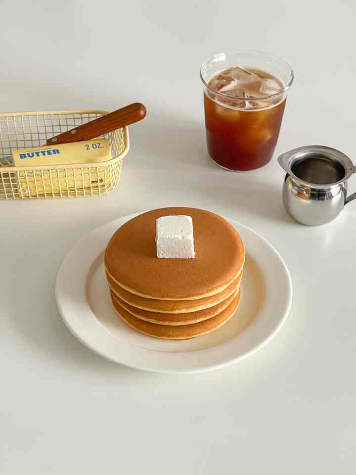 白いテーブルの上に積まれた茶色いパンケーキとバター、アイスティーが置かれたナチュラルな雰囲気のおうちカフェ