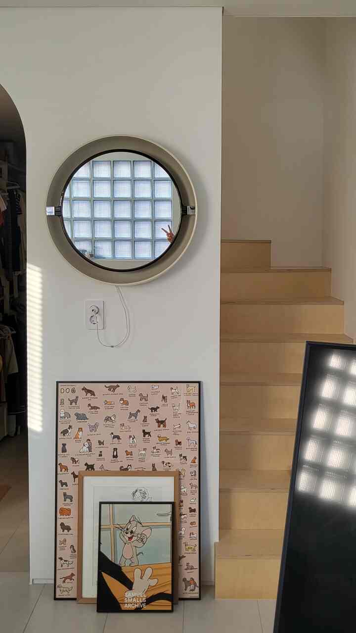 白い壁とナチュラルな木製階段の玄関空間、丸い鏡と複数のポスターが配置されたすっきりとしたインテリア