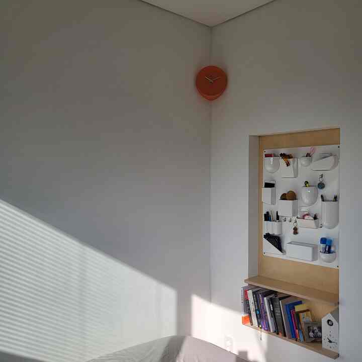白を基調とした寝室、小さな本棚と壁掛けの収納ボックス、そして壁に設置されたカッコークロックがある雰囲気のあるインテリア