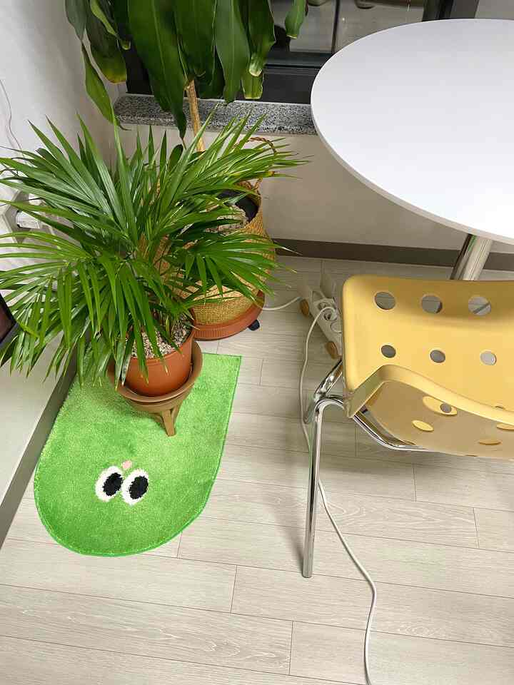 ホワイトの床とダイニングテーブル、イエローのダイニングチェアが配置された小さな空間に、緑の植物と遊び心のあるグリーンラグがあるナチュラルなインテリア