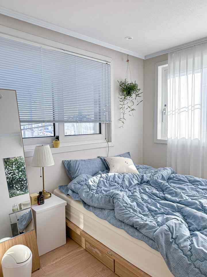 白と青のトーンの寝室、大きなベッドと枕、窓辺のブラインドと植物が配置された落ち着いた雰囲気のインテリア