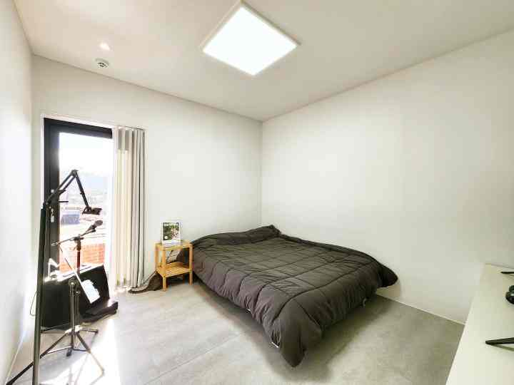 白い壁と天井が広がる一人暮らし向け寝室、黒い寝具とナイトテーブル、遮光カーテンが配置されたシンプルな空間
