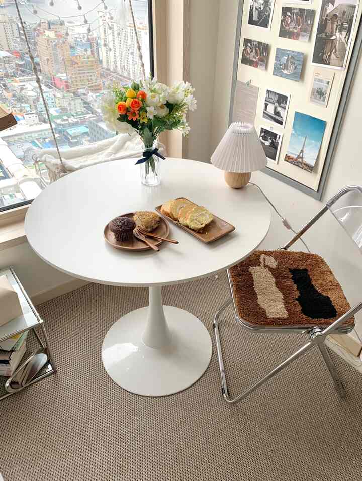 白とベージュの色調のダイニングルーム、円形テーブルと折りたたみ椅子、壁に飾られたポスターがある雰囲気のある空間