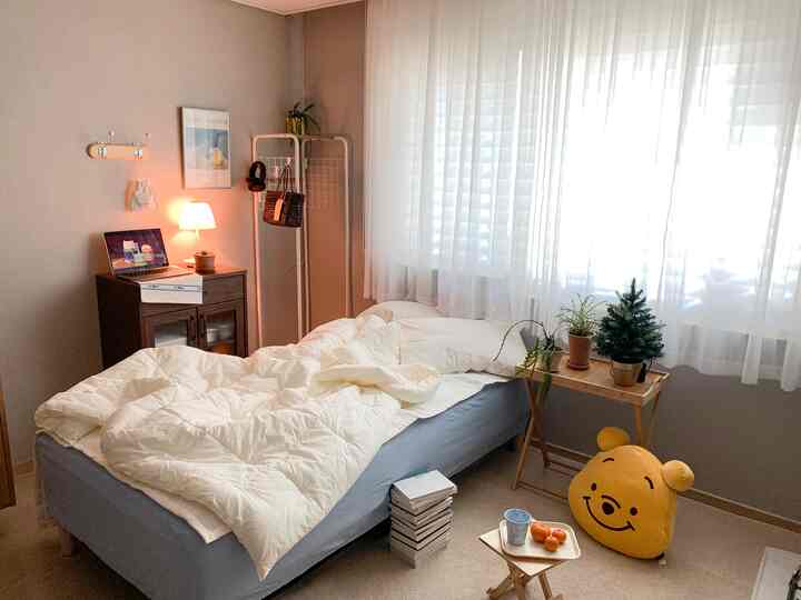 ホワイトとブラウンのトーンによる6畳サイズのシンプルでナチュラルな寝室、ベッドやナイトテーブル、ハンギングラック、植物が配置された落ち着いた雰囲気の空間