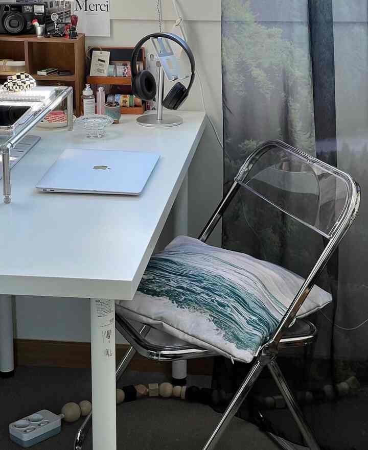 ホワイトトーンのコンパクトなホームオフィス、透明な折りたたみ椅子と白いデスクにノートパソコンとヘッドホンが置かれた整った作業空間