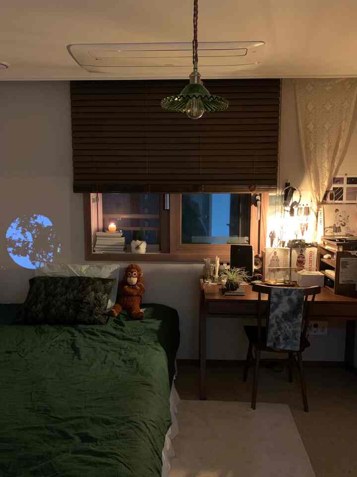 暗めの色調の寝室、濃い茶色のブラインドと緑色のベッドカバー、木製のデスクとペンダントライトが配置された雰囲気のある空間